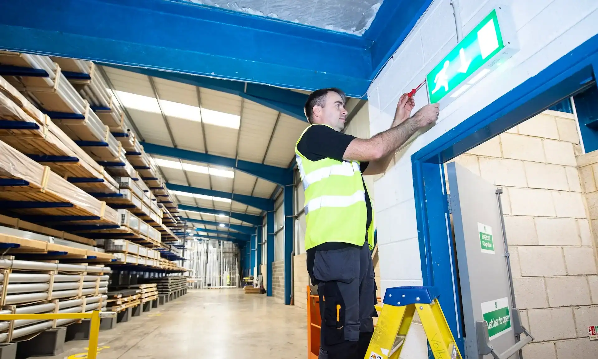 Electrician in a high-visibility vest inspecting an illuminated emergency exit light above a doorway in an industrial warehouse.
